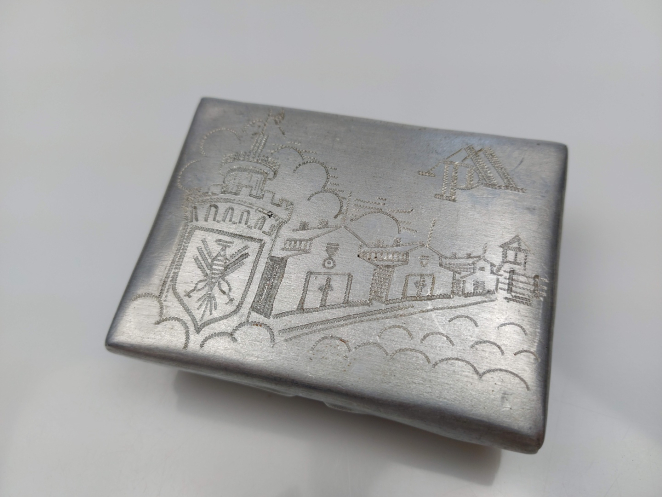 Metalowa papierośnica, na niej, z lewej strony wygrawerowany herb, z prawej fragment zabudowań obozu.