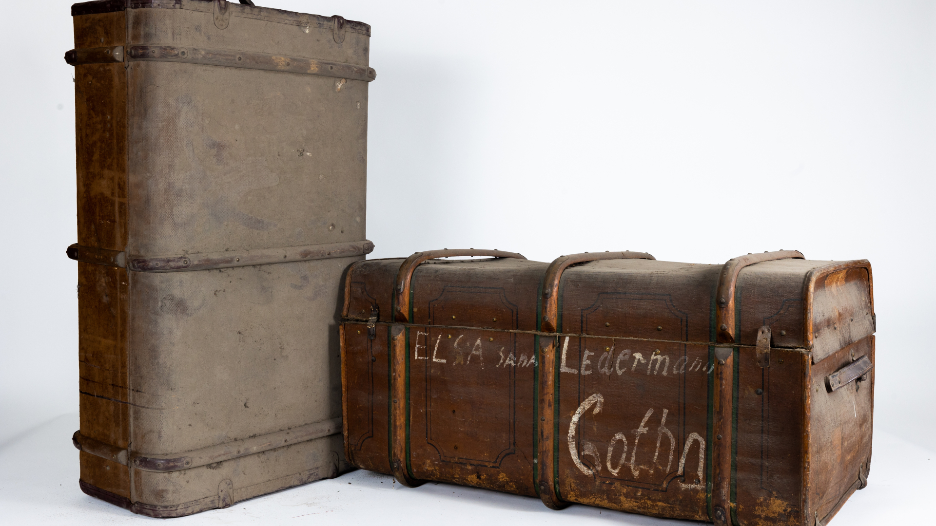 Dwie stare, brązowe walizki, z prawej stoi w pionie, lewa w poziomie, wieko zamknięte, poniżej na walizce białe napisy. 