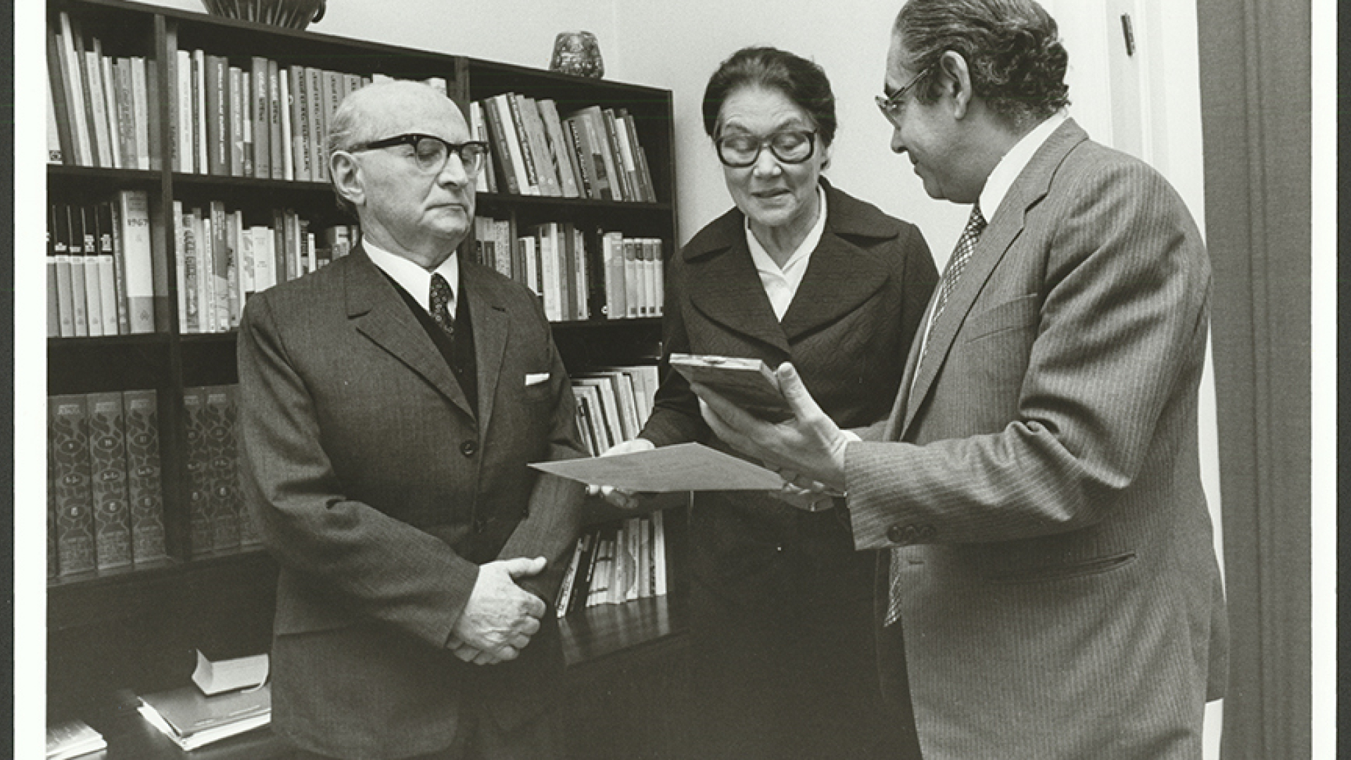 Czarno-białe zdjęcie, w środku kobieta, po dwóch stronach mężczyźni w garniturach. Kobieta trzyma w ręce biała kartkę, mężczyzna po lewej niewielką odznaczenie, w tle biblioteczka.