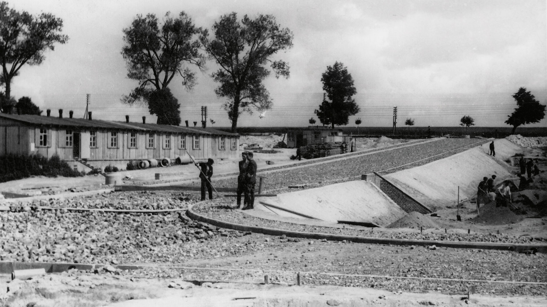 Fotografia czarno-biała, historyczna, przedstawiająca teren budowy, na środku trzech mężczyzn, jeden kopie w ziemi łopatą, z lewej strony w tle budynek, za nim drzewa 