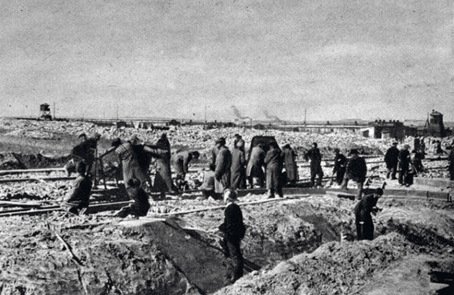 Grupa kilkunastu osób ubranych w pasiaki i stroje robocze, trzymających łopaty, dookoła hałdy ziemi. 