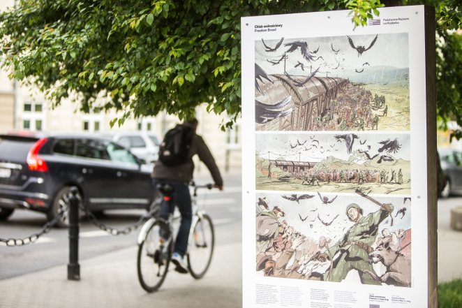 Panel wystawy przestawiającej stronę komiksu Chleb wolnościowy, na drugim planie rowerzysta oraz jadący samochód