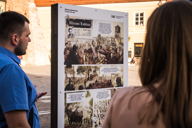 Mężczyzna w niebieskiej koszuli obok panelu wystawienniczego z przedstawioną kartą komiksu