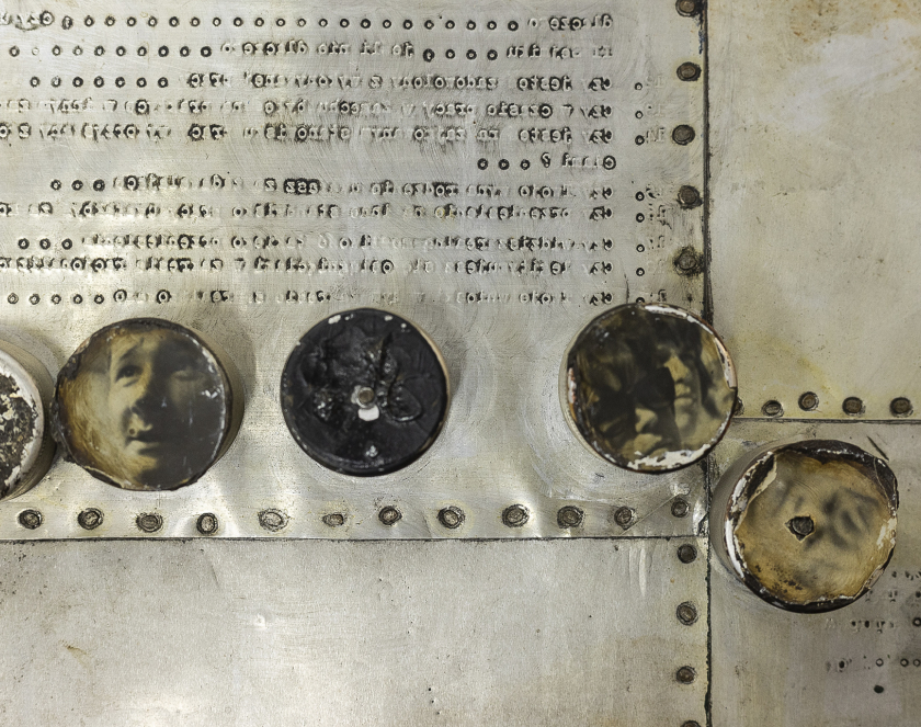 metalowe blaszki połączone za pomocą metalowych nitów, widoczne cztery metalowe tonda z niewyraźnymi fotografiami, na jednej powiększona twarz mężczyzny 