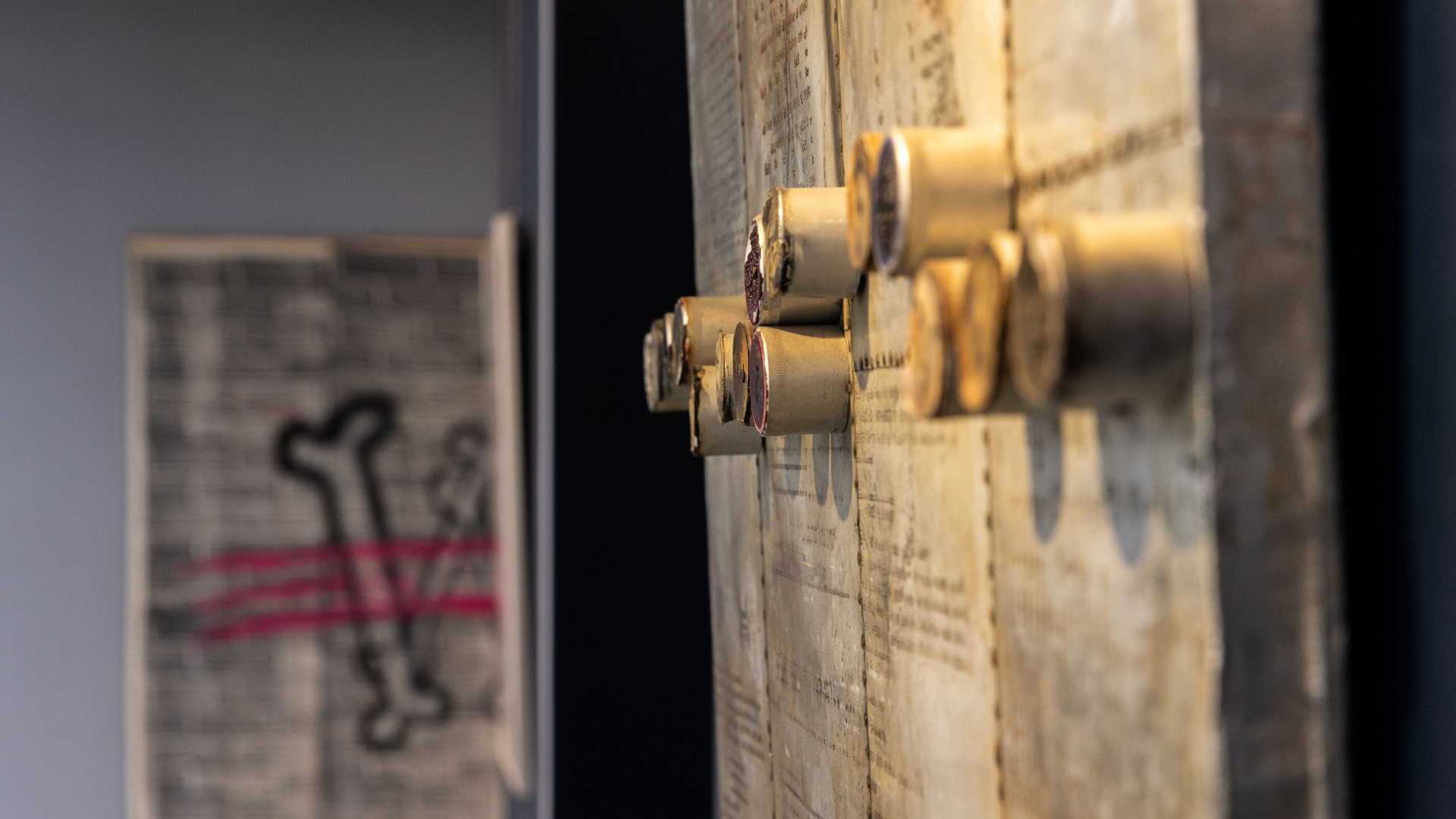 Widok na wystawę, z prawej strony w zbliżeniu praca Mariana Bogusza, w formie metalowej płyty, w tle fragment pracy Tomka Kawiaka, widać literę W z piszczeli przekreśloną różową farbą