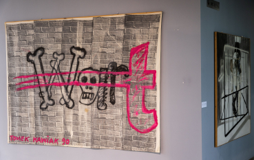 Dwa obrazy zawieszone na ścianach, pierwszy wyraźny: słowo WAR(t), war stworzone z kości oraz czaszki w kolorze czarnym, t - jaskrawy różowy, rozmazany pasmami poziomymi przez cały obraz, w tle mur złożony z szarych cegieł, na dole obrazu napis: Tomek Kawiak 90