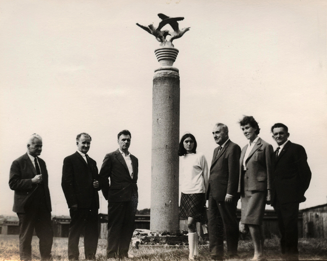 Pięciu mężczyzny w średnim wieku ubranych w garnitury, dwie kobiety (jedna starsza, druga młoda), obok kolumna Trzech Orłów