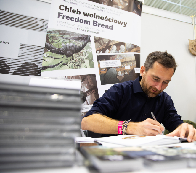 40-letni mężczyzna podpisujący książkę, w tle baner z napisem: Chleb wolnościowy, Freedom bread 