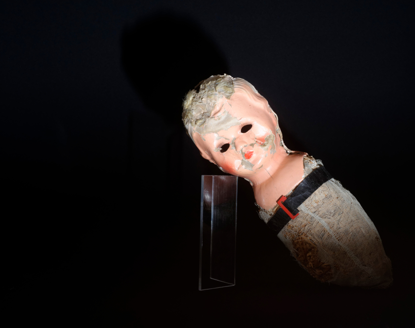 Zniszczona głowa i korpus lalki wsparta o plastikowy postument, dookoła czarne tło