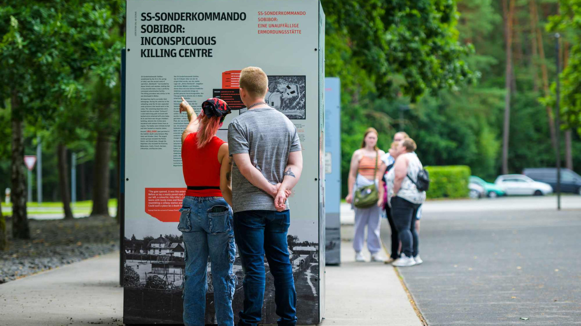 Zdjęcie współczesne, na chodniku przy ulicy stoi wystawa plenerowa, dwoje ludzi, kobieta i mężczyzna, ustawionych tyłem do fotografa ogląda tablice wystawy, kobieta wskazuje ręką na tekst. W tle grupa osób. 
