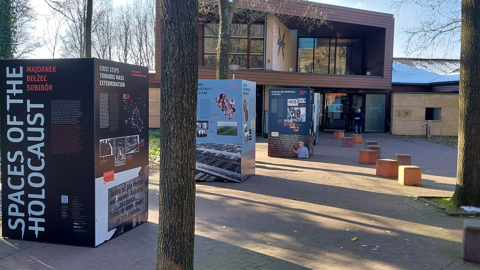 Zdjęcie współczesne, kolorowe, po lewej stronie wystawa plenerowa, w tle nowoczesny, przeszklony budynek muzeum.