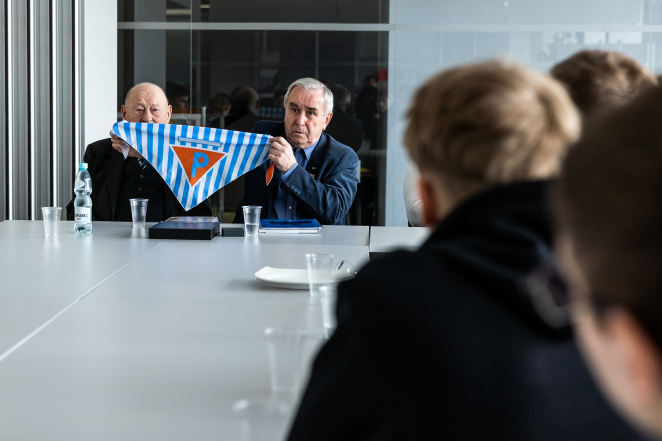 Współczesne zdjęcie kolorowe. Dwóch mężczyzn pokazuje chustę w niebiesko-białe pasy, na środku odwrócony trójkąt z literą P. Z prawej strony młodzież, siedzą skierowani w stronę mężczyzn