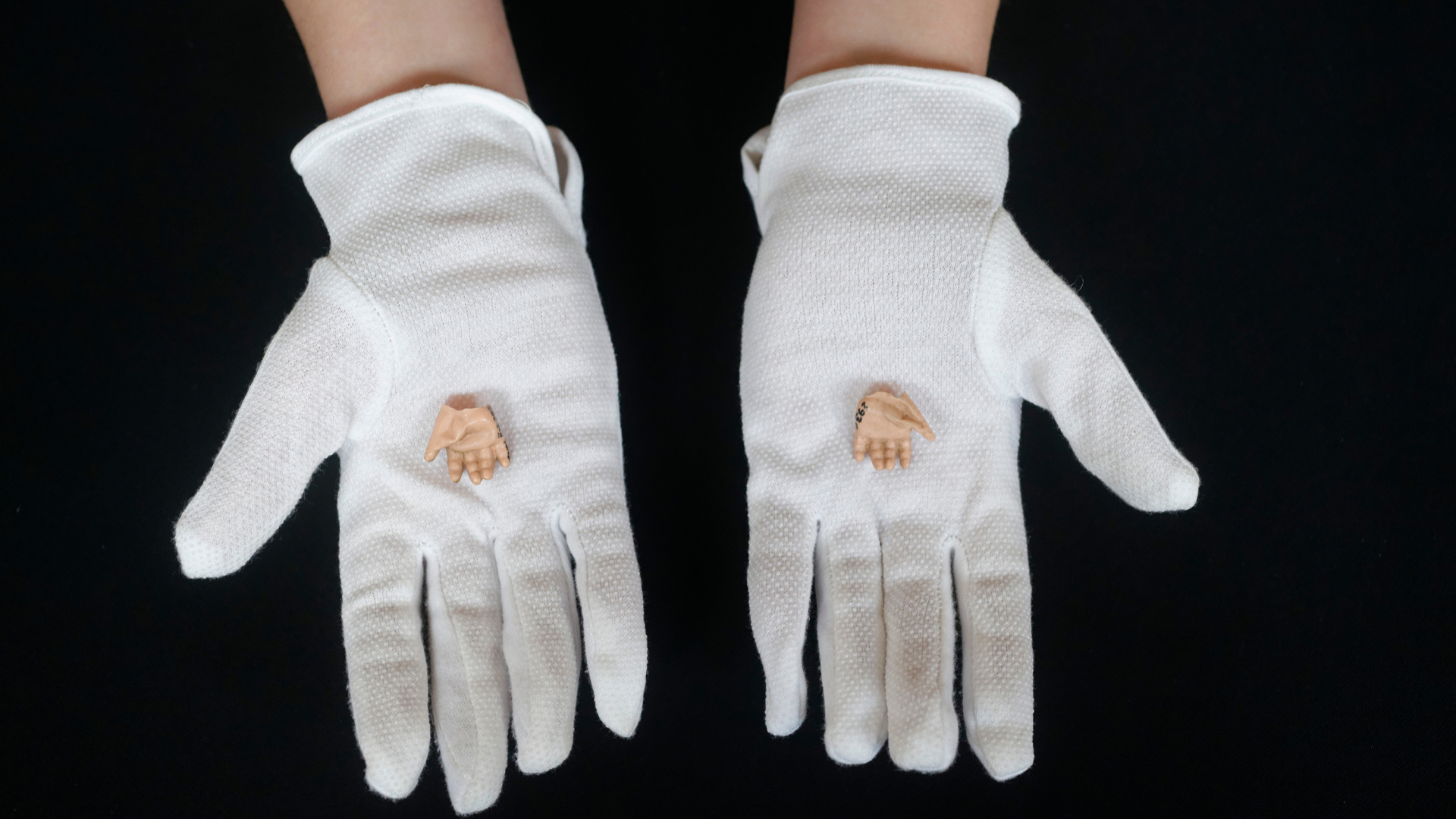 Dwie dłonie pokazane kciukami do zewnątrz, w białych rękawiczkach, na nich ułożone dłonie lalek, tło czarne 