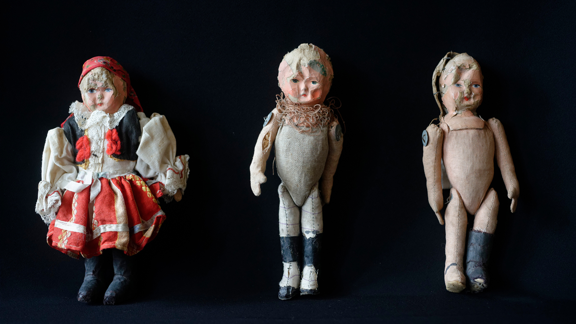 Trzy lalki na czarnym tle, z lewej w ubraniu ludowym, dwie bez ubranek, twarze wgniecione 