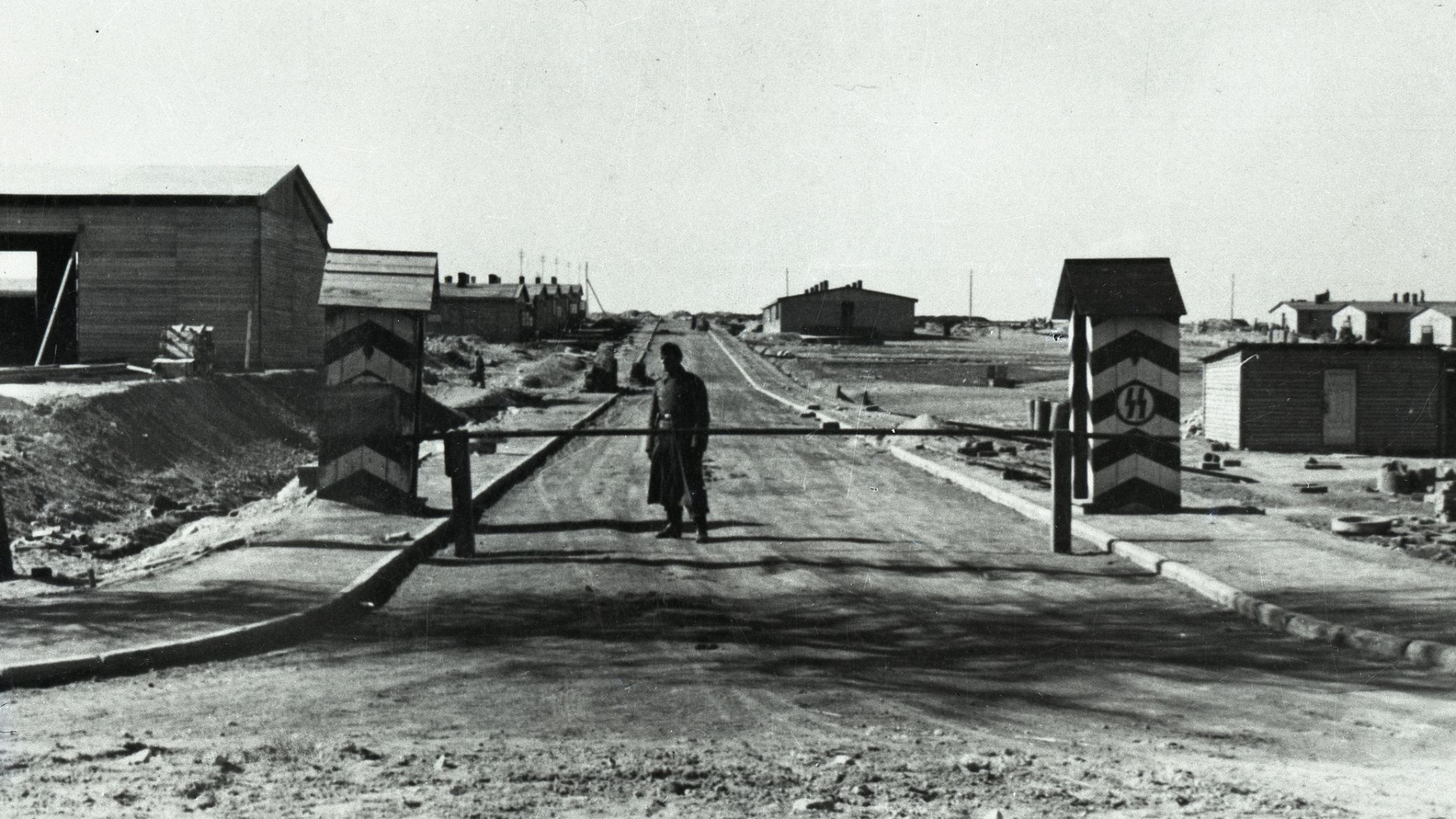 Czarno-białe zdjęcie, droga, szlaban, dwie budki wartownicze z symbolami nazistowskimi, porośrodku mężczyzna w mundurze. 
