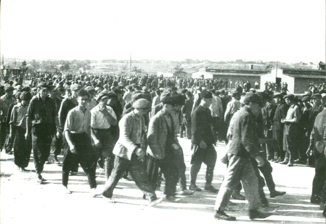 Na pierwszym planie grupa kilkunastu idących młodych mężczyzn. W tle dwa baraki oraz grupa kilkudziesięciu mężczyzn