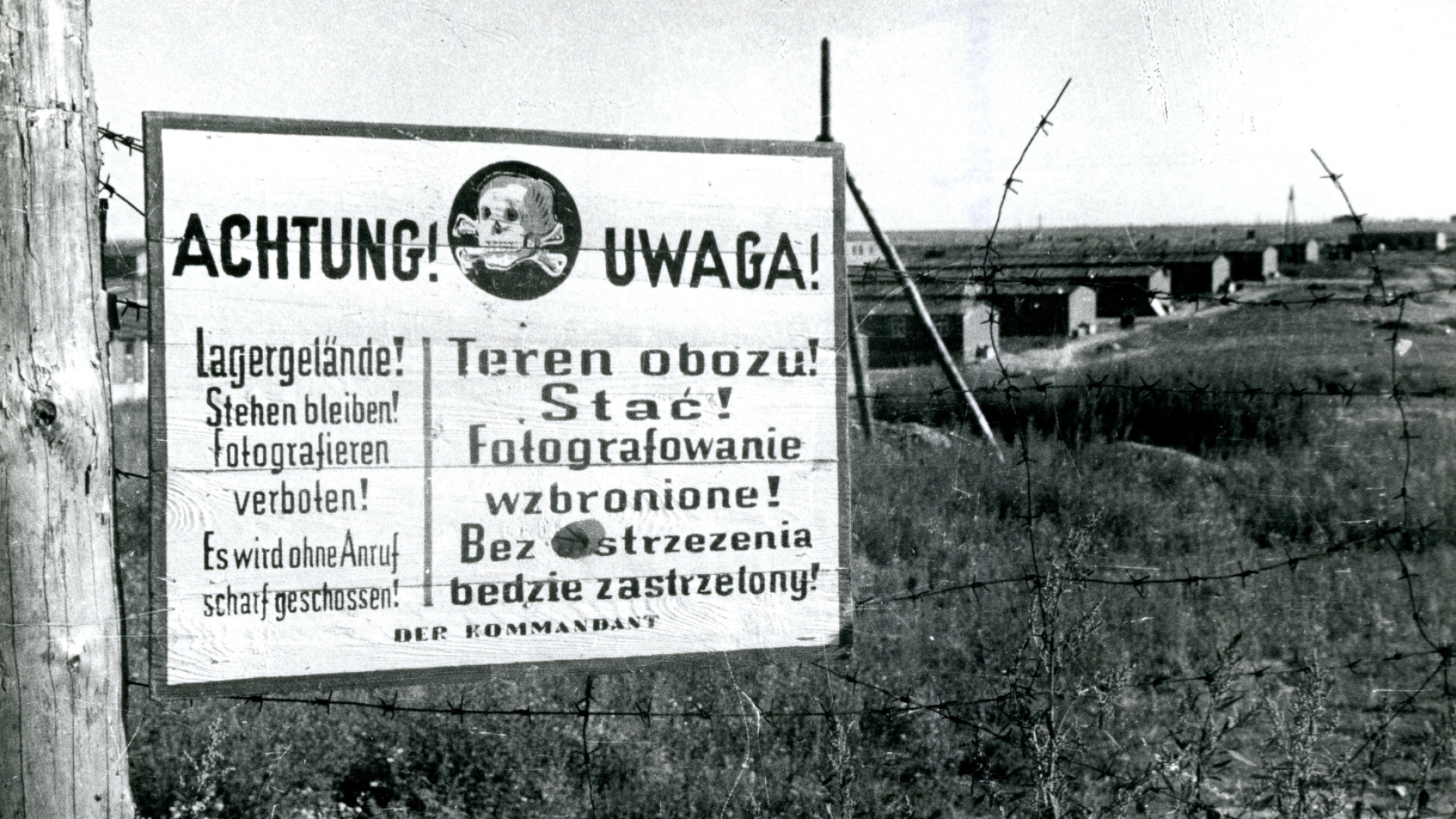 Czarno-białe zdjęcie, na pierwszym planie biała tablica w języku polskim i niemieckim z ikoną czaszki i dwóch krzyżowanych kości piszczelowych, w te rząd drewnianych baraków więźniarskich. 