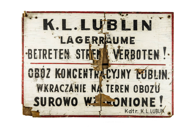 Biała metalowa tablica z napisem w języku polskim i niemieckim o treści: Obóz koncentracyjny Lublin. Wkraczanie na teren obozu surowo wzbronione!