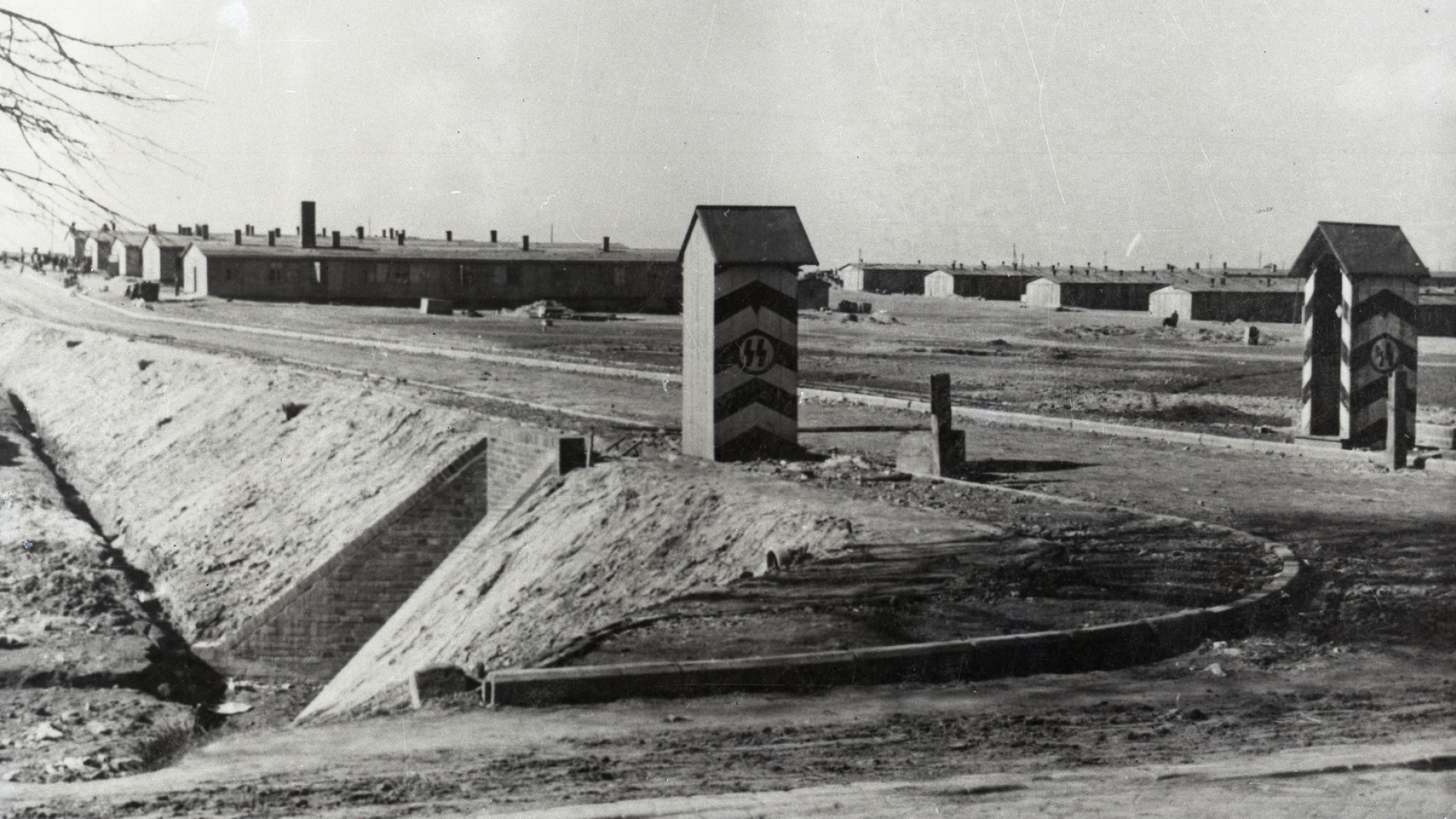 Czarno-białe zdjęcie, droga, po obu jej stronach budki wartownicze z nazistowskimi symbolami, w tle baraki więźniarskie. 