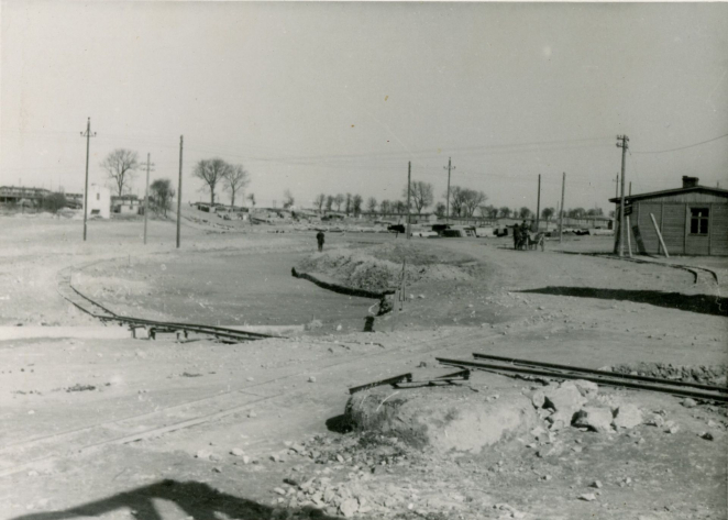 Uklepana ziemia, na której leżą szyny. W tle sylwetki pojedynczych osób. Po prawej fragment parterowego baraku  