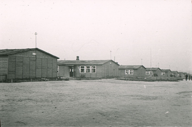 Rzędy drewnianych piętrowych baraków. Po prawej stronie widoczne sylwetki ludzi