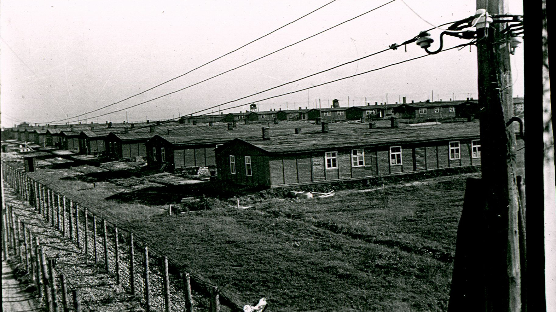 czarno-białe zdjęcie, rzędy baraków więźniarskich, na pierwszym planie podwójne ogrodzenie z drutu kolczastego. 