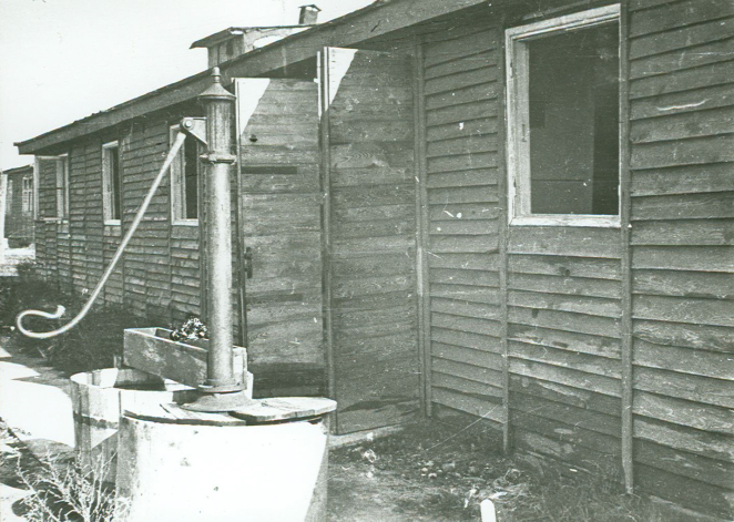 Drewniany budynek z otwartymi drzwiami. Przed nim studnia z metalową pompą