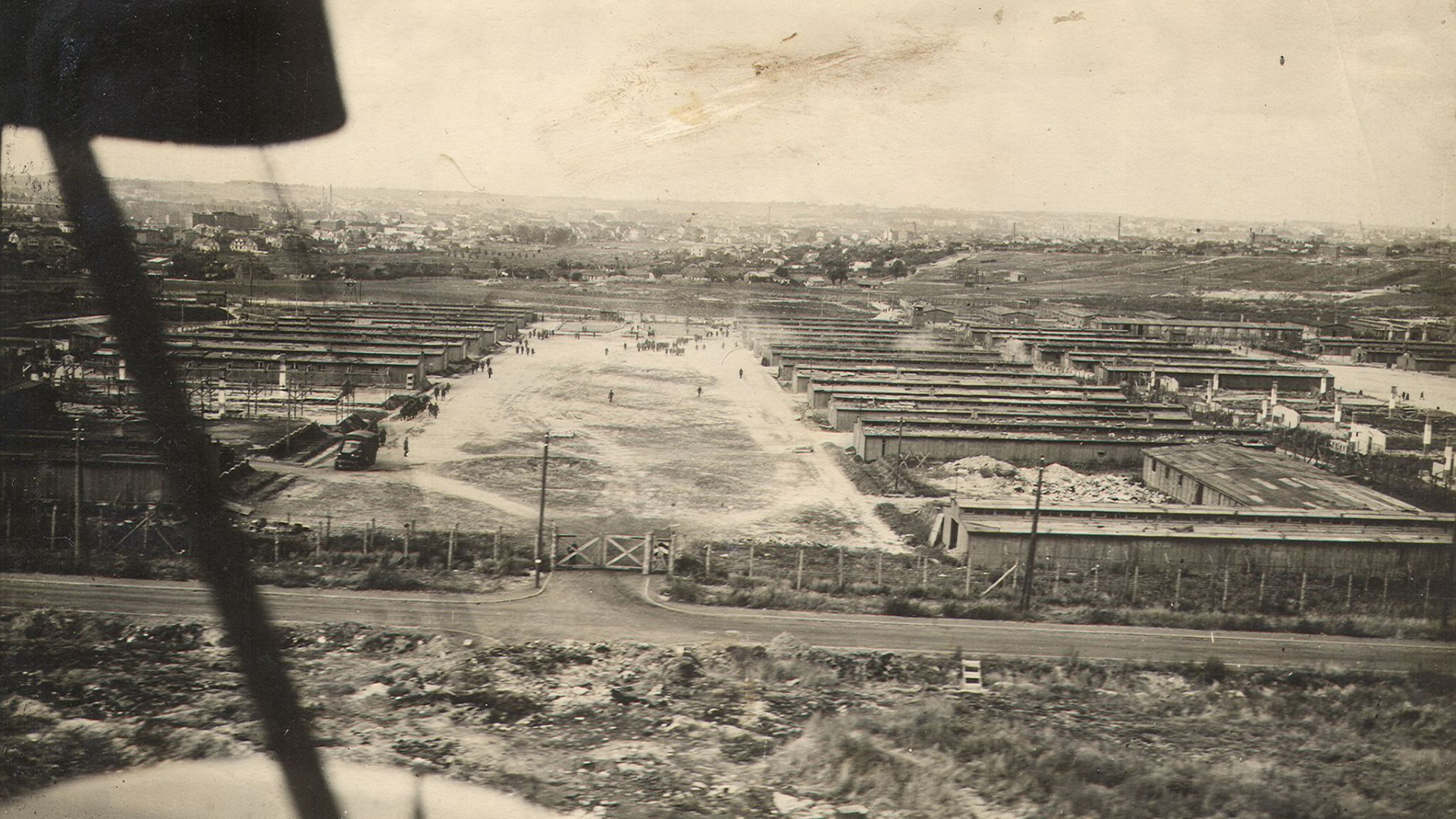 Zdjęcie w septii wykonane z lecącego samolotu, przestrzeń ogrodzona drutem kolczastym, pośrodku klepisko dookoła rzędy drewnianych baraków 