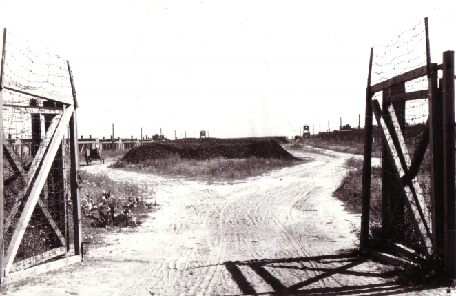 Otwarta brama z drewna i drutu kolczastego. Za nią nasyp ziemny. Po jego bokach niskie budynki drewniane