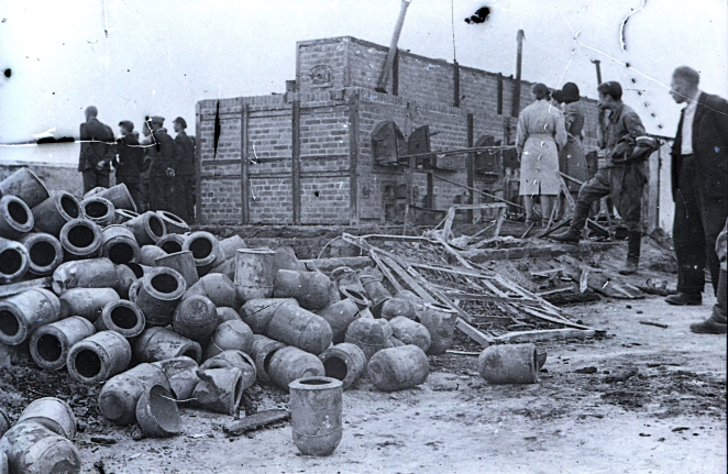 Piece z cegły. Przed nimi stos urn. Dookoła ludzie przyglądający się pozostałościom po krematorium.