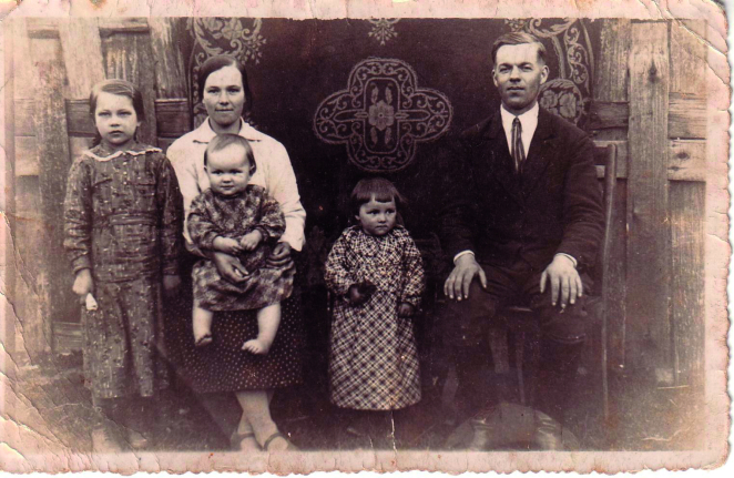 Na zdjęciu siedzą dwie dorosłe osoby - kobieta i mężczyzna. Między nimi stoi mała dziewczynka w kraciastej sukience. U kobiety na kolanach siedzi druga mała dziewczynka w kraciastej sukience. Obok niej stoi trzecia dziewczynka w wieku około 10 lat w długiej sukience z kołnierzykiem