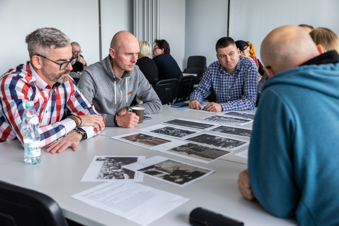 Grupa mężczyzn w średnim wieku siedzi przy stole, na środku czarno białe wydruki zdjęć historycznych 
