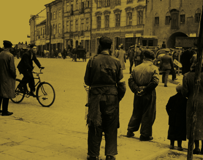 Grupa osób stojąca na ulicy, jeden mężczyzna jadący na rowerze, w tle miejskie zabudowania