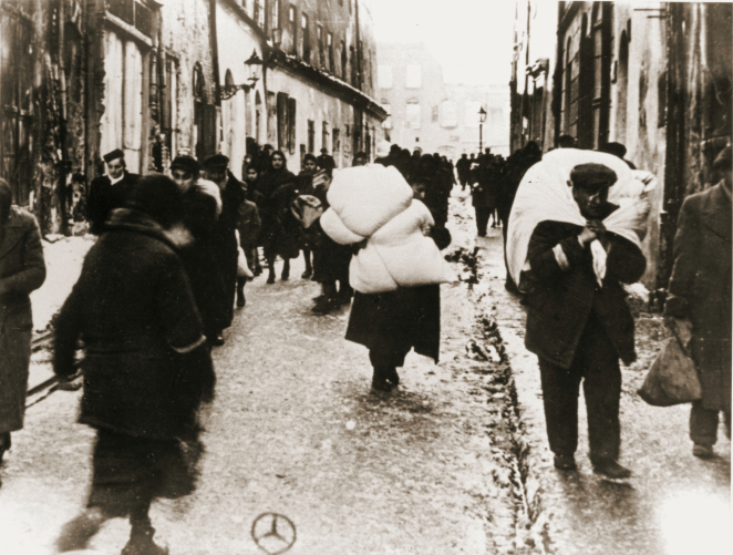 Miejska ulica, grupa osób idąca pośród kamienic, dwóch mężczyzn niesie tobołki