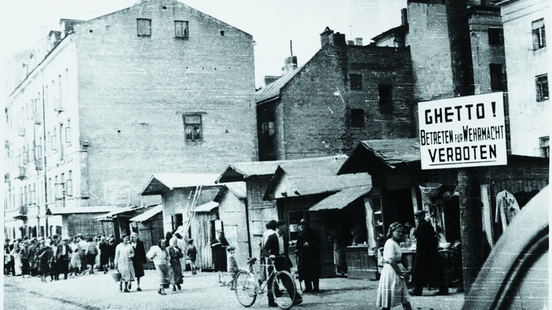 Zdjęcie czarno-białe, historyczne, miejskie zabudowania, wzdłuż nich ulicą idą ludzie, na pierwszym planie kobieta w sukience, w centrum mężczyzna prowadzi rower, po lewej stronie tablica z napisem: Getto! Betreten fur Wermacht Verboten  