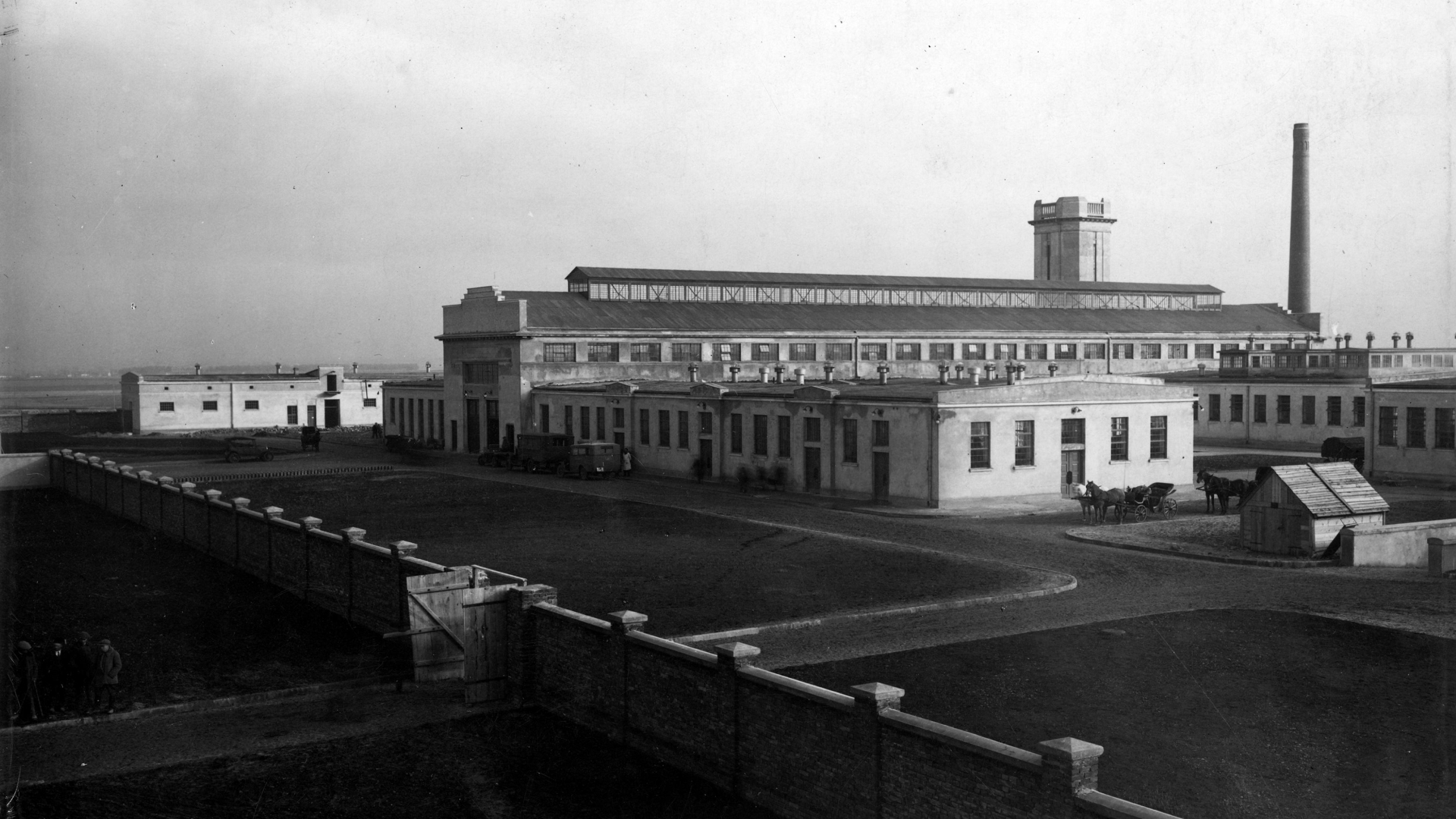 Zdjęcie czarno-białe, historyczne, na pierwszym planie, ukośnie ogrodzenie z cegły z wejściem, na drugim okazały budynek z dużą ilością okien