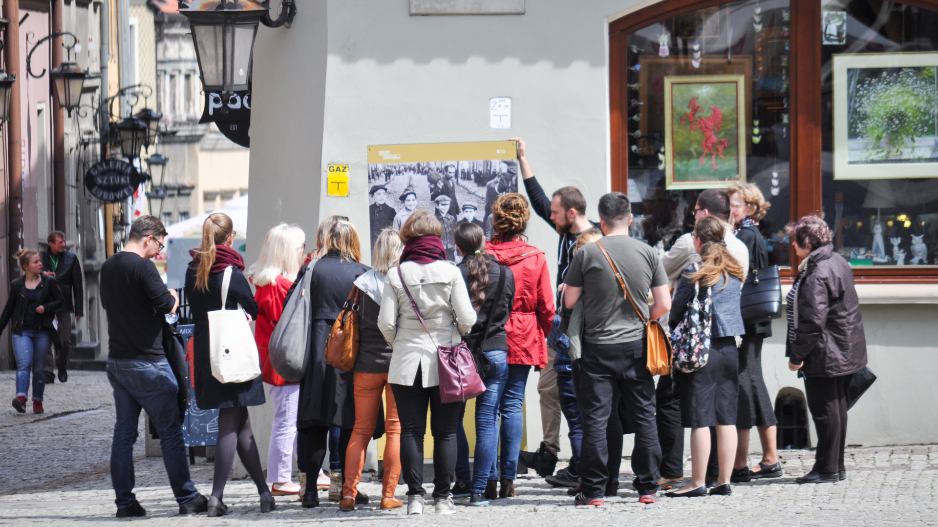 Duża grupa ludzi stoi przy tablicy wystawienniczej ustawionej w przestrzeni Starego Miasta