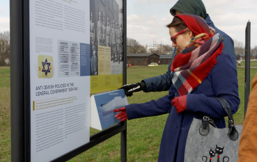 Kobieta w kolorowej chuście, czerwonych rękawiczkach i okularach przeciwsłonecznych przy obrotowym elemencie panelu wystawienniczego wystawy plenerowej, na drugim planie baraki więźniarskie