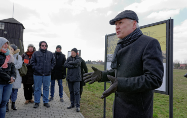 50-letni mężczyzna ubrany w ciemny płaszcz i ciemno szary kaszkiet stoi na tle panelu wystawy, widoczna grupa osób i wieża wartownicza  