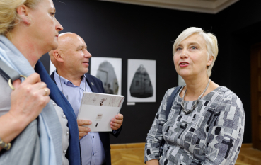 50-letnia kobieta o jasnych krótkich włosach rozmawia z dwójką osób, mężczyzna trzyma katalog wystawy 