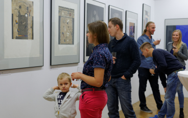 Grupa osób oglądająca prezentowane na wystawie dzieła, na pierwszym planie kilkuletni chłopiec obok kobiety w czerwonych spodniach 