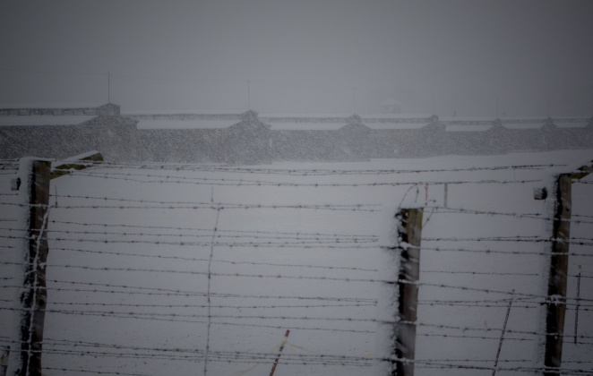 Rząd baraków więźniarskich podczas zamieci śnieżnej, na pierwszym planie ogrodzenie z drutu kolczastego.