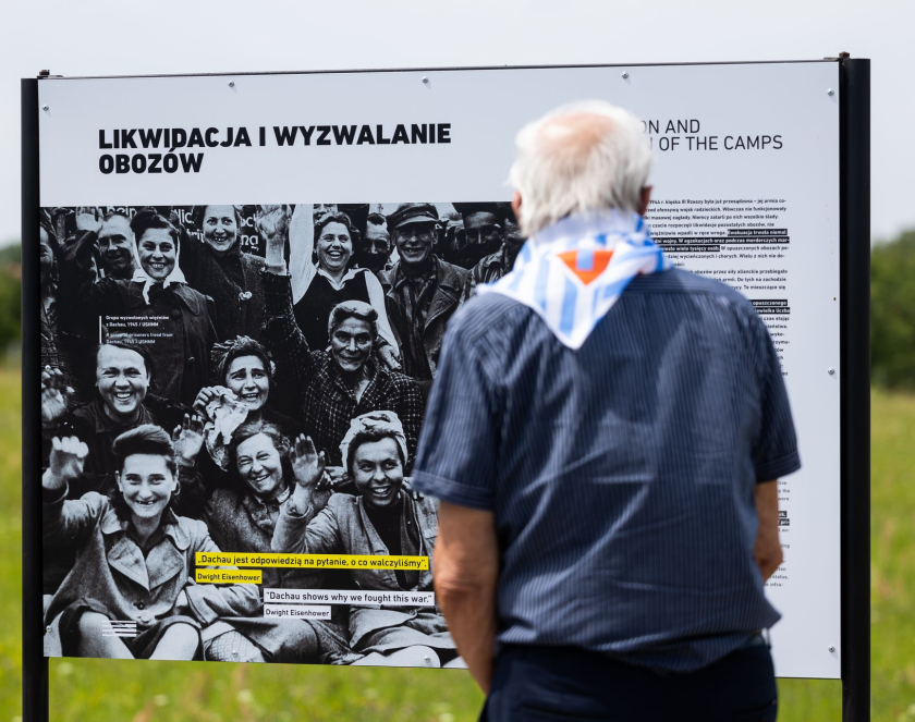Starszy mężczyzna z chustą w biało-niebieskie paski i czerwony trójkąt z literą P czyta treść wystawy plenerowej, napis na planszy:  Likwidacja i wyzwalanie obozów 