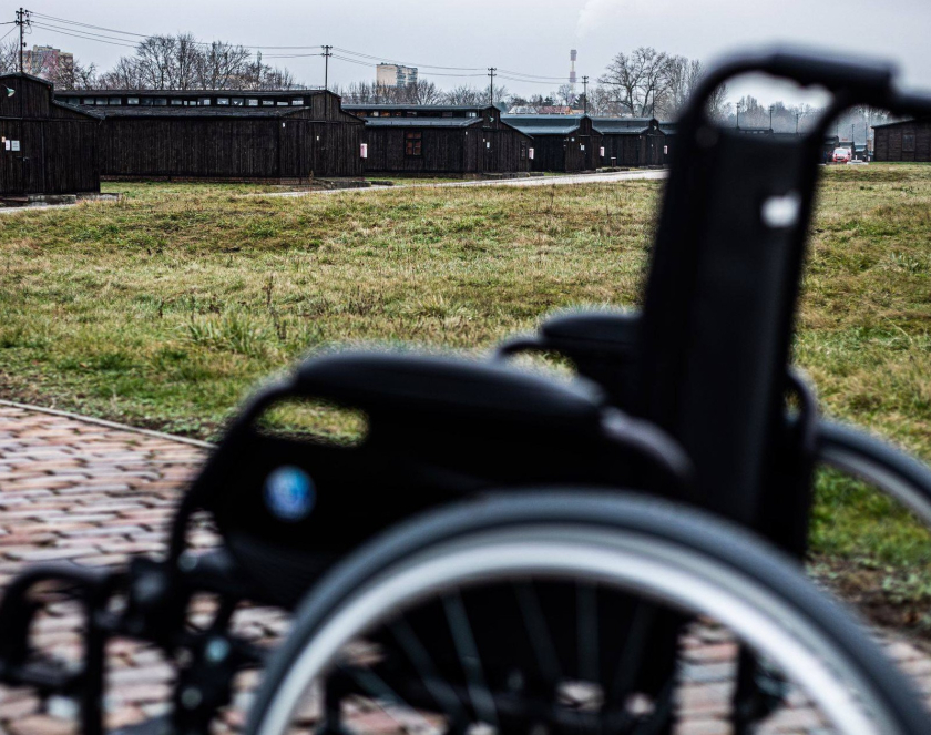 Wózek inwalidzki w tle rząd baraków więźniarskich. 