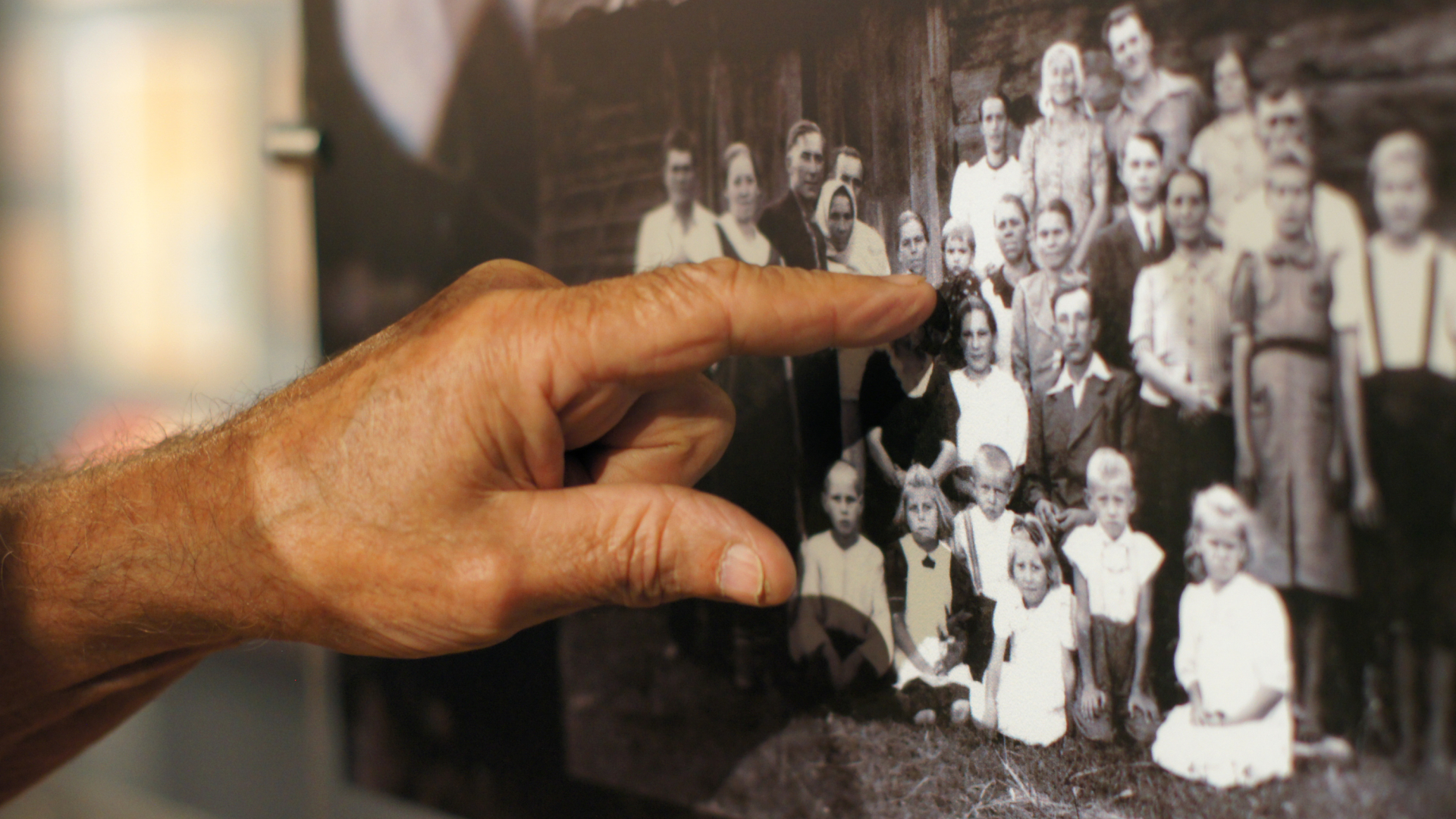 Dłoń starszej osoby wskazuje na czarno-białą fotografię portretową kilkudziesięciu osób