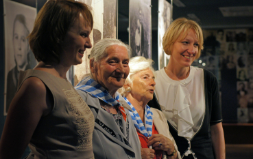 Cztery uśmiechnięte kobiety na tle wystawy, dwie starsze z chustami w biało niebieskie pasy