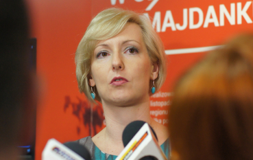 Młoda kobieta o blond włosach na tle banneru z napisem: Wysiedleńcy z Zamojszczyzny w obozie na Majdanku 