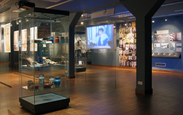 Wnętrze wystawy przeszkolone gabloty z przedmiotami, panel multimedialny z relacją wideo, w tle historyczne zdjęcia oraz portrety 