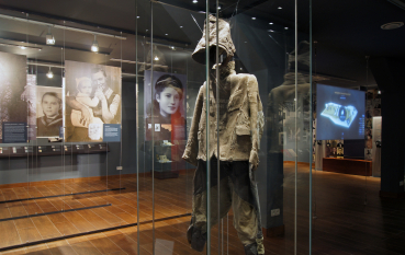 Wnętrze wystawy, porwane ubranie dziecięce w szklanej gablocie, na drugim planie wielkoformatowe zdjęcia 