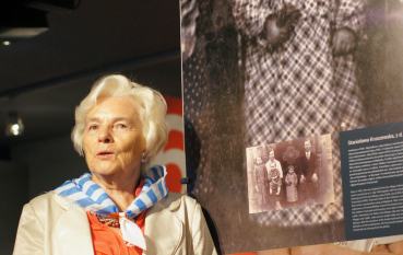 Starsza kobieta ubrana w białą marynarkę z chustą w biało niebieskie pasy stoi pod wielkoformatowym zdjęciem małej dziewczynki ubranej w kraciastą sukienkę 